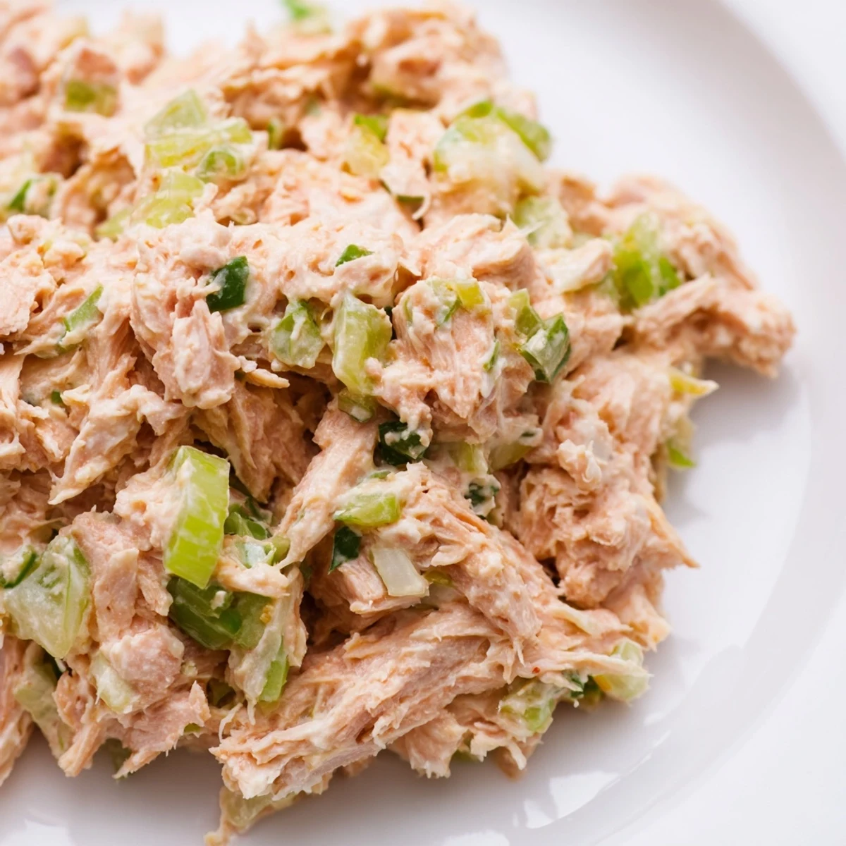 A close-up of Spicy Zesty Tuna Salad in a white bowl, featuring chunks of tuna, vibrant green scallions, and a creamy, orange-hued dressing garnished with fresh parsley.  